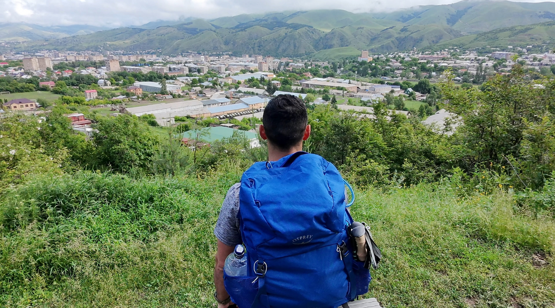 Edmond Margaryan with a blue Osprey backpack sits overlooking the city of Vanadzor, Armenia, surrounded by lush green mountains and valleys - the starting point of an epic walking journey from Vanadzor to Dubai.