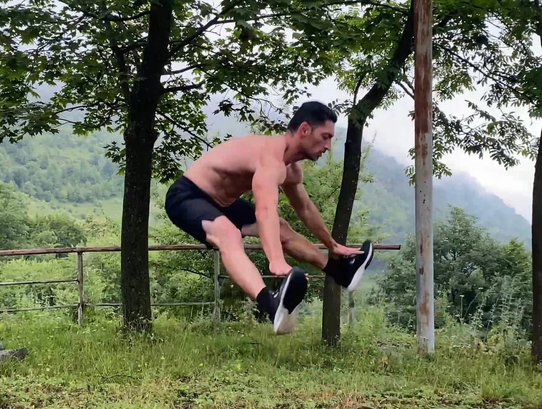 Edmond Margaryan mid-air performing an explosive burpee variation in an outdoor forest setting, demonstrating strength, balance, and body control against a lush green mountain backdrop—symbolizing raw fitness and mental grit.